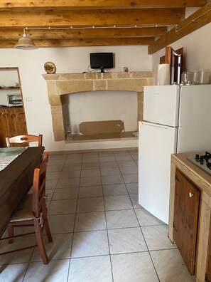 Een keuken met een stenen open haard, een televisie erboven, een houten eettafel met stoelen en een koelkast.