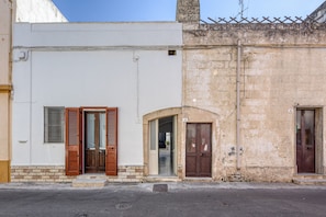 Un edificio con fachada blanca, contraventanas de madera en la planta baja y un muro de piedra a la derecha.