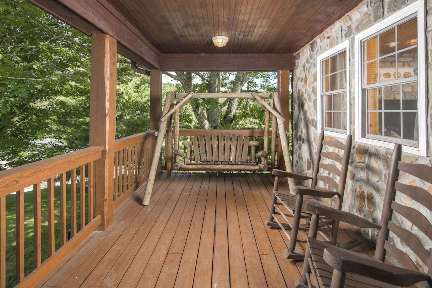 A wooden deck with a swing, stone walls, and a view of trees.