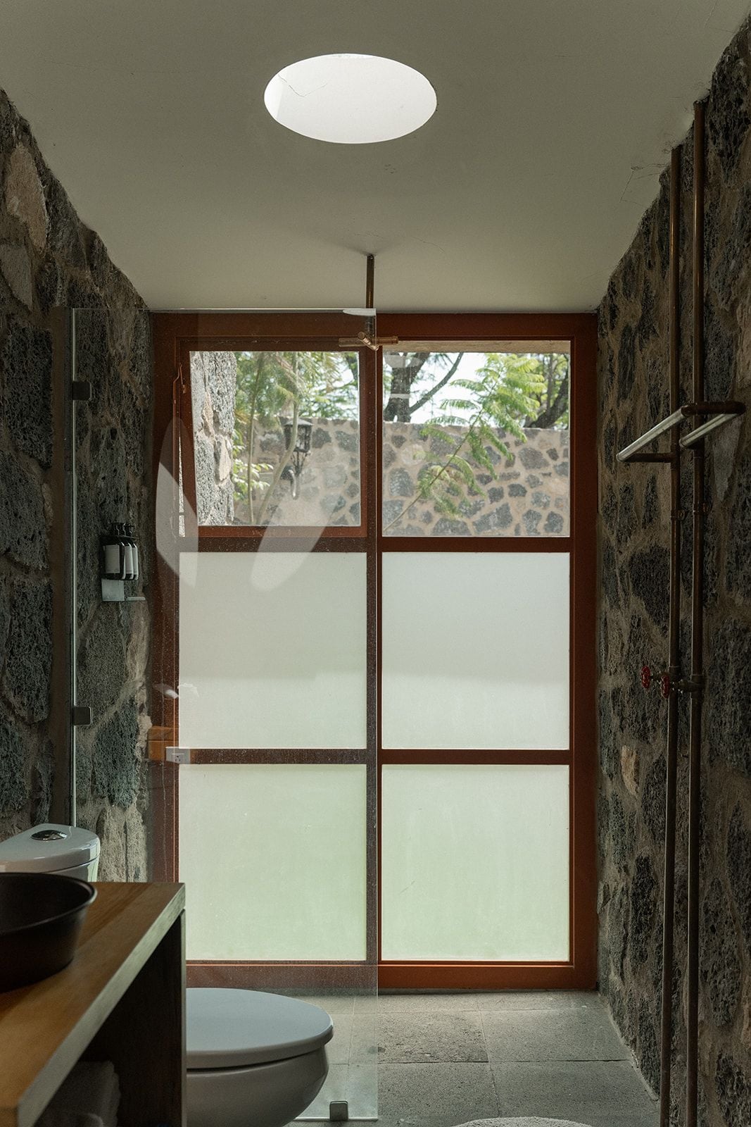 Une salle de bain avec un mur en pierre, des toilettes, un lavabo et une fenêtre donnant sur des arbres.