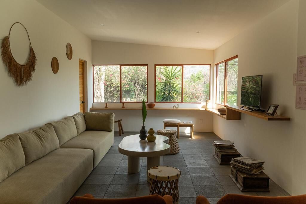 A living room with a sofa, a round table, a television, and a window with a view of greenery.