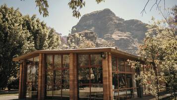 Un pavillon entièrement vitré, avec une base en briques, entouré d’arbres et adossé à une montagne rocheuse.