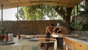 An outdoor kitchen with a brick oven, wooden table, chairs, and potted plants.