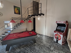 Una mesa de billar con fieltro rojo, una vara de billar y una lámpara roja. Hay una máquina expendedora de Coca-Cola y una escalera con barandilla de metal.