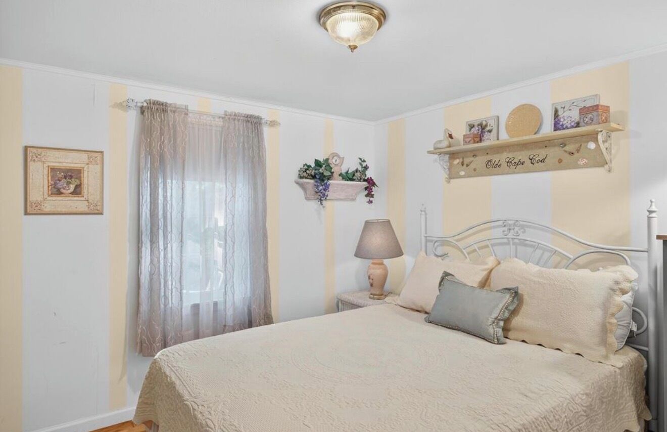 Une chambre à coucher comprenant un lit, une table de chevet, une lampe, une fenêtre avec des rideaux, une étagère fixée au mur avec des objets décoratifs et un tableau encadré accroché au mur.