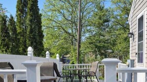 A wooden deck with a white railing, outdoor furniture, and a view of trees and a building. - Relaxing Cape Cod Cottage Getaway (Dennis)
