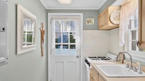 A kitchen with a white door, a stove, a sink, and wooden cabinets. - Relaxing Cape Cod Cottage Getaway (Dennis)