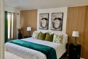 Ein Schlafzimmer mit einem Bett, zwei an der Wand befestigten Bildern, einem Nachttisch, einer Lampe und einem Fenster mit Vorhängen.