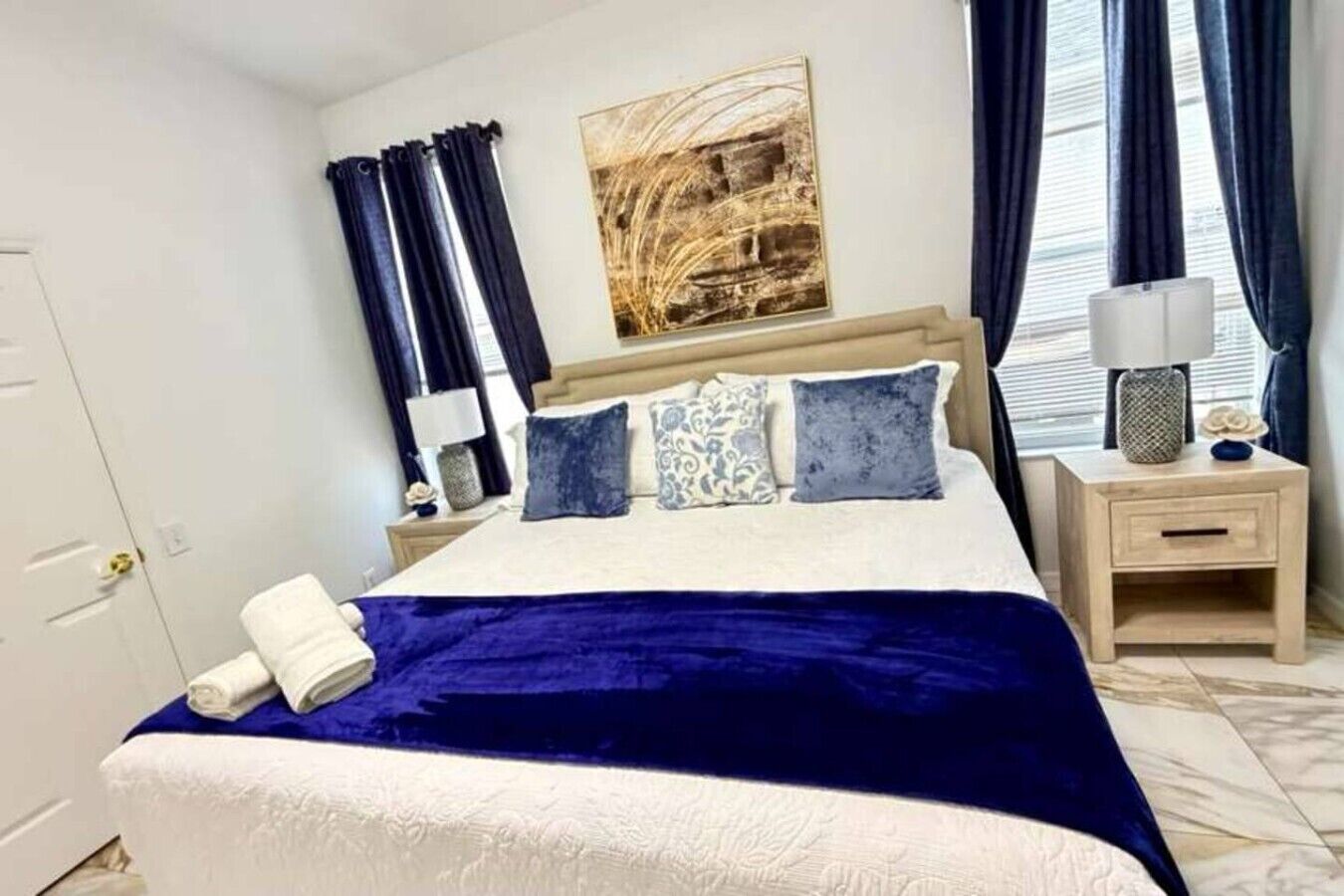 Une chambre à coucher avec un lit, une table de chevet, une lampe, une fenêtre avec des rideaux et un tableau au mur.