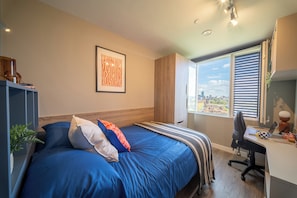 Ein Schlafzimmer mit Bett, Schreibtisch, einem Fenster mit Jalousien, einem gerahmten Bild an der Wand und Blick auf die Stadt.