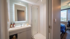 Une salle de bain moderne dotée d’une douche à parois de verre, d’un meuble-lavabo blanc avec des articles de toilette, et offrant une vue sur la ville par une fenêtre.