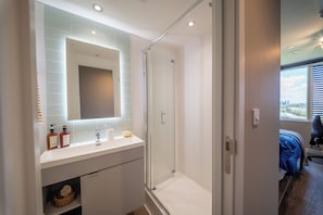 A modern bathroom with a glass shower enclosure, a white vanity with toiletries, and a view of the city through a window. - Ironworks House (London)