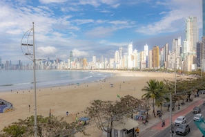 A beachside cityscape with high-rise buildings, a sandy beach, and a road with vehicles. - Apto de luxo com piscina privativa e vista mar (Balneário Camboriú)