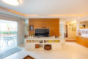 A modern living room with a flat-screen TV, wooden wall paneling, a dining area with chairs, and a kitchen with a glass display case. - Apto de luxo com piscina privativa e vista mar (Balneário Camboriú)