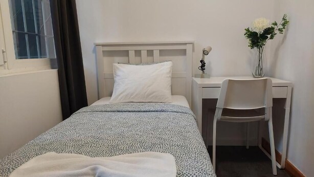 Una cama individual con colcha estampada, una mesita de noche con lámpara, una silla y un florero con flores sobre un escritorio.