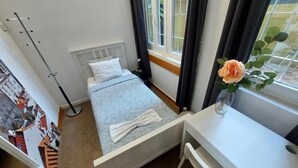Habitación pequeña con cama, escritorio, ventana con cortinas y vistas a edificios.