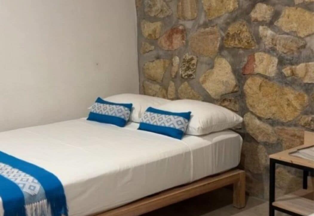 Una cama individual con estructura de madera, ropa de cama blanca y almohadas estampadas en azul, frente a una pared de piedra.