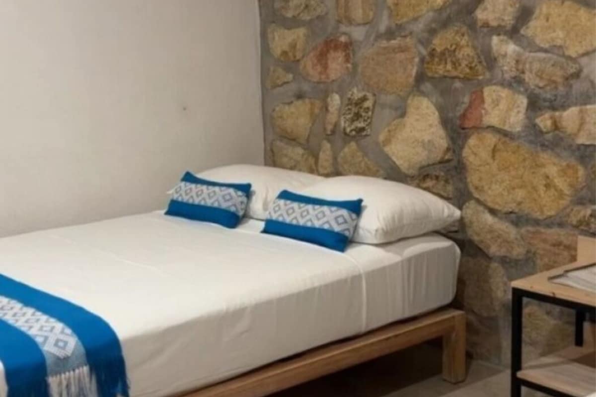 Uma cama de solteiro com estrutura de madeira, roupa de cama branca e almofadas com padrão azul em frente a uma parede de pedra.