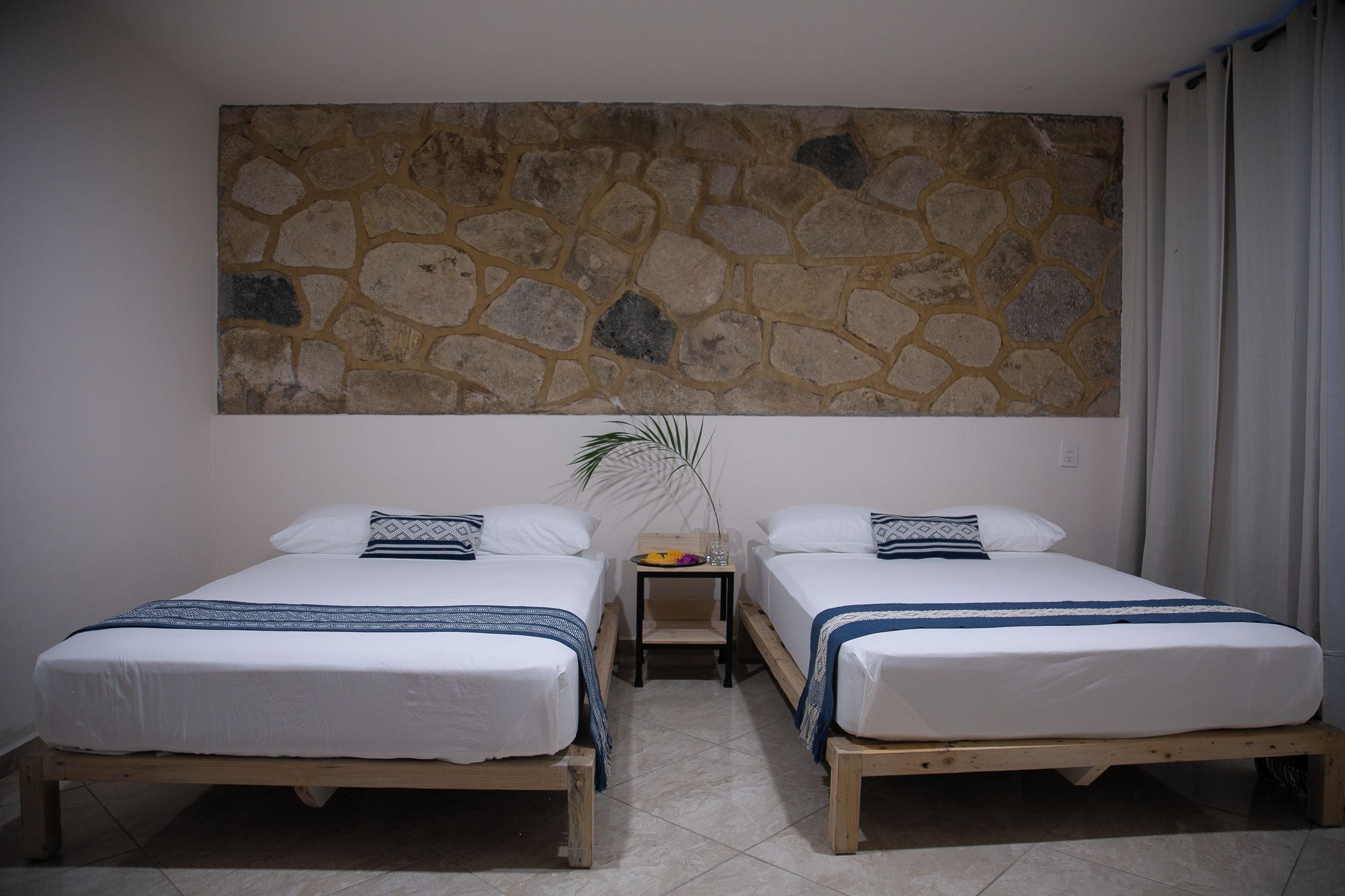 Una habitación con dos camas de madera, una pared de piedra, una mesita con una planta y una ventana con cortinas.