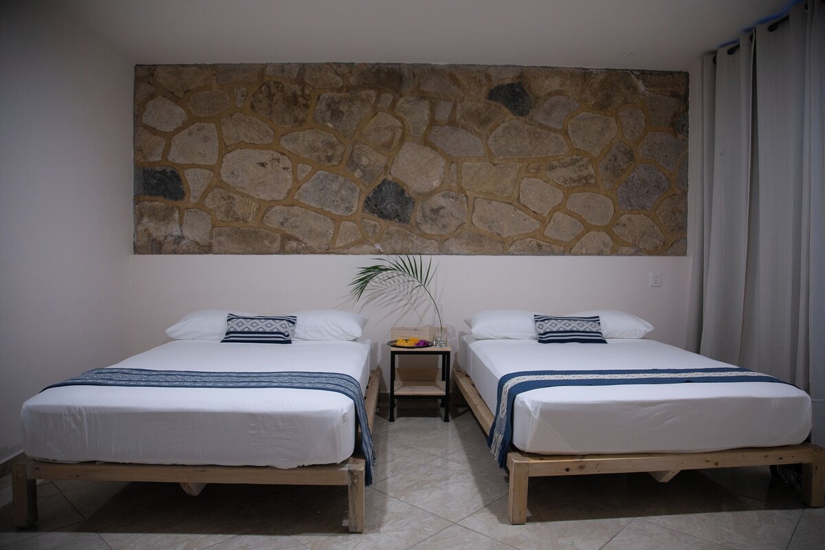 A room with two wooden beds, a stone wall, a small table with a plant, and a window with curtains.