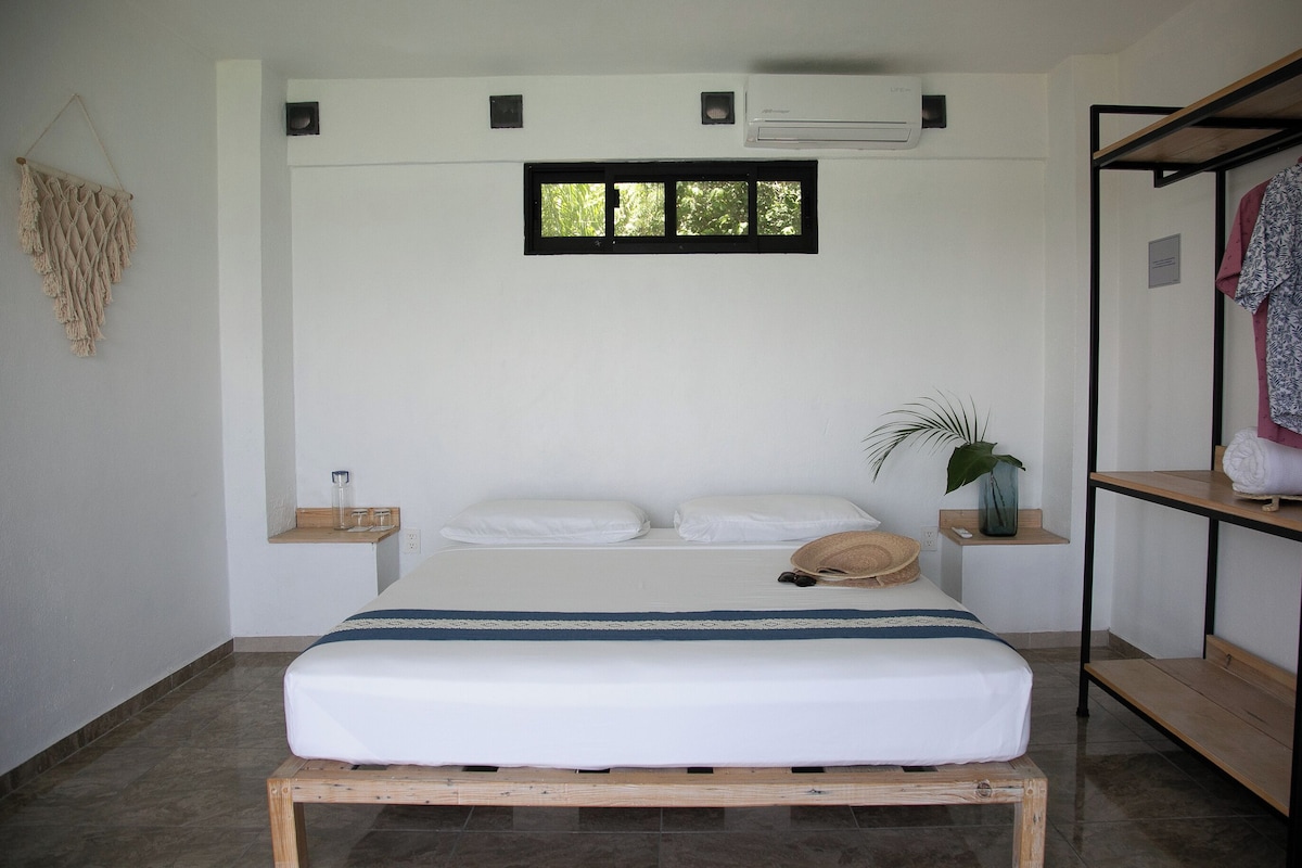 Quarto com cama, janela, prateleira com objetos e um pendente de macramé na parede.
