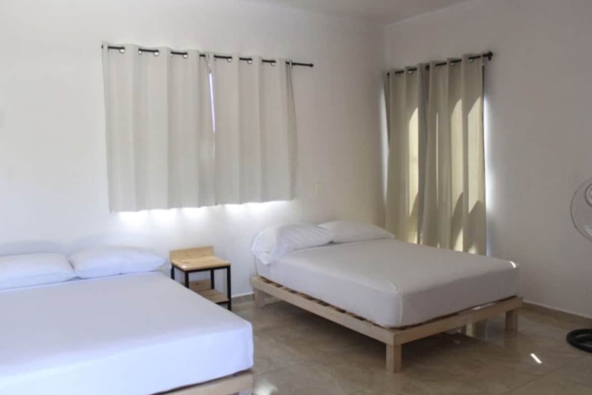 Quarto com duas camas, uma mesinha e uma janela com cortinas.