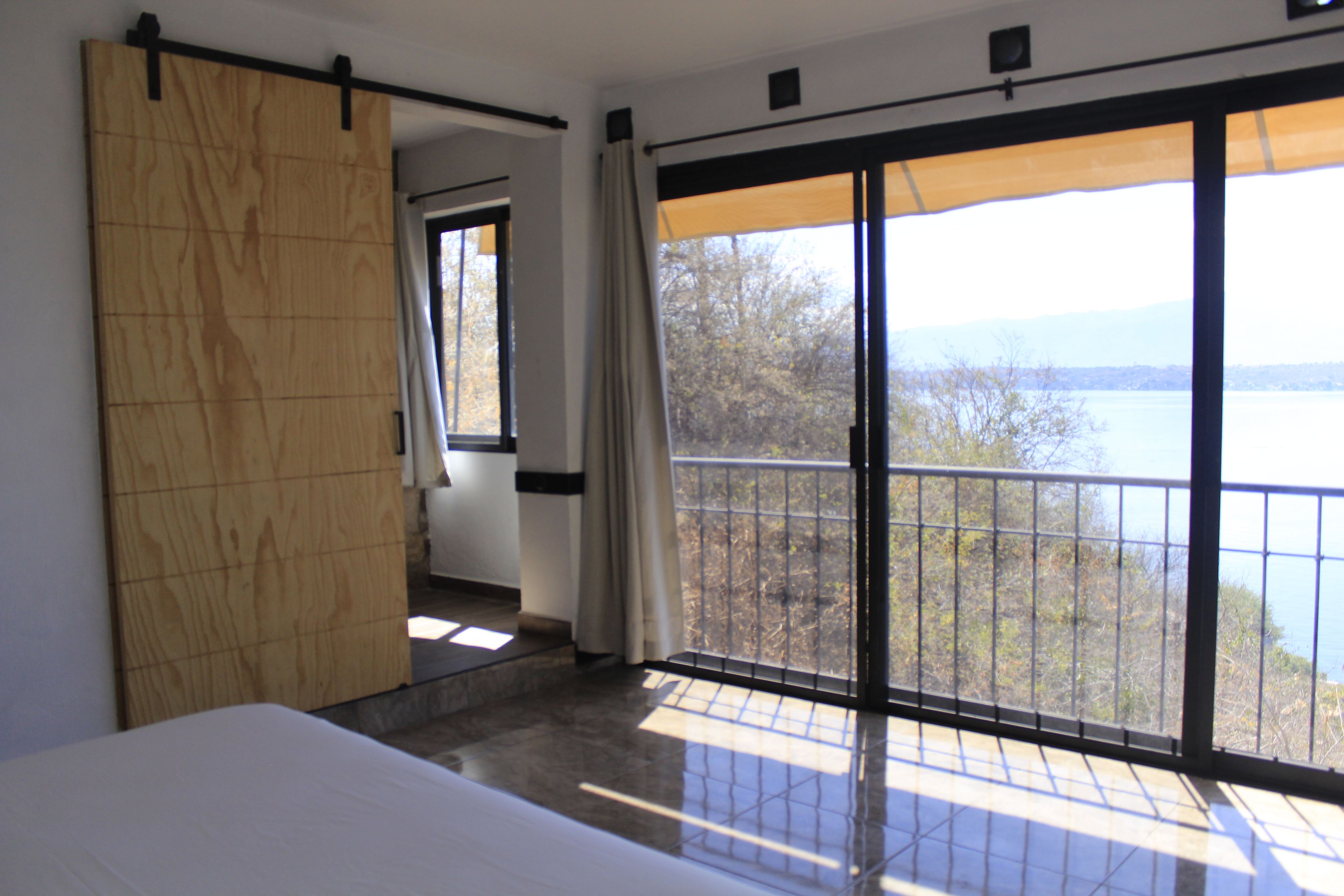 Un dormitorio con un amplio ventanal corredero que da a un balcón con vista a un lago y árboles.