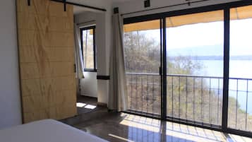 Un dormitorio con un amplio ventanal corredero que da a un balcón con vista a un lago y árboles.