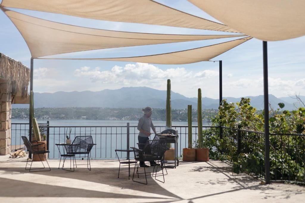 Un patio exterior con vista a montañas y una masa de agua, que incluye una mesa, sillas, una parrilla y una persona encargándose de la parrilla.