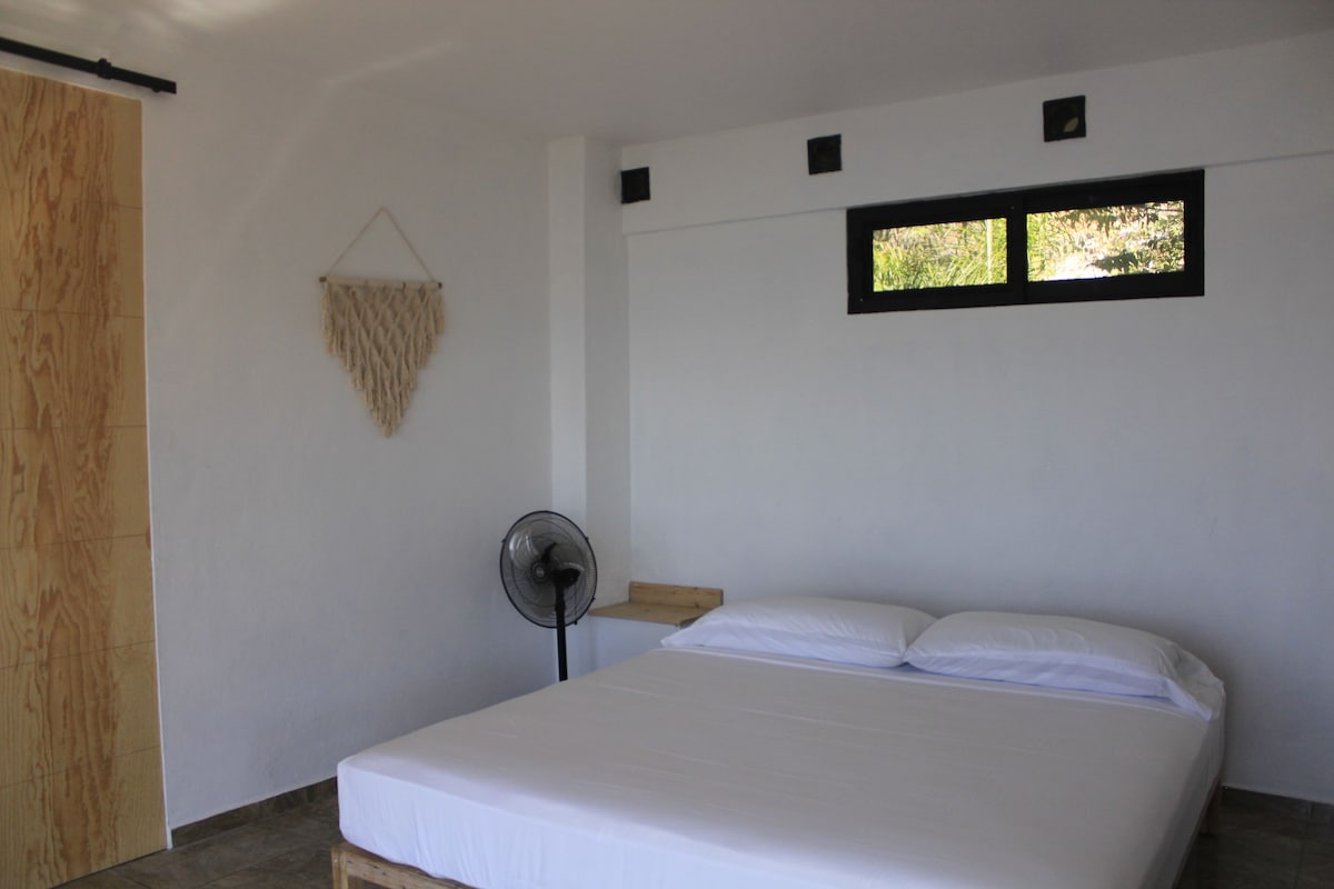 A bedroom with a bed, a wall-mounted fan, a small window, and a wooden door.