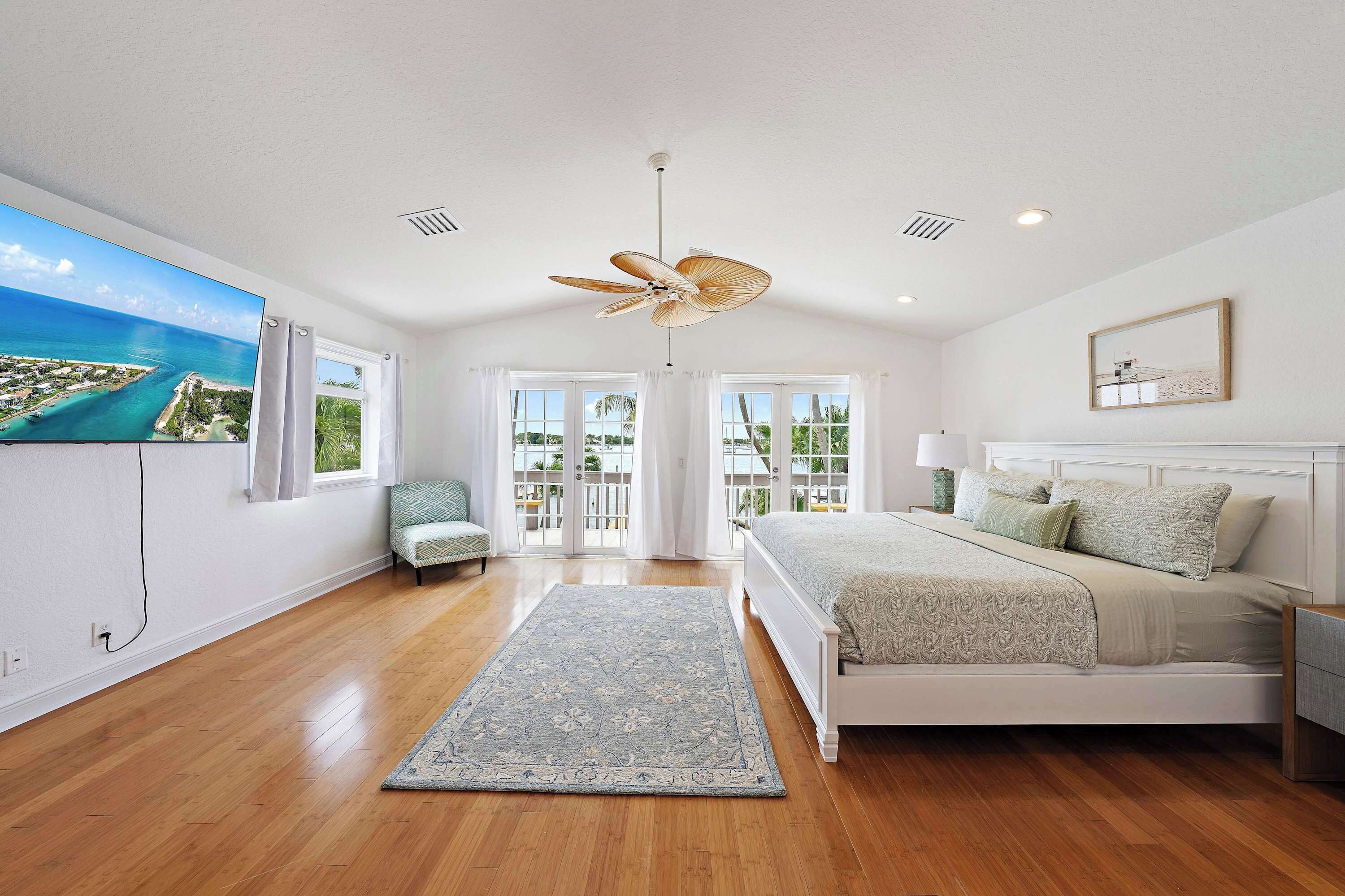 A bedroom with a large bed, a ceiling fan, a view of the ocean, and a window with curtains.