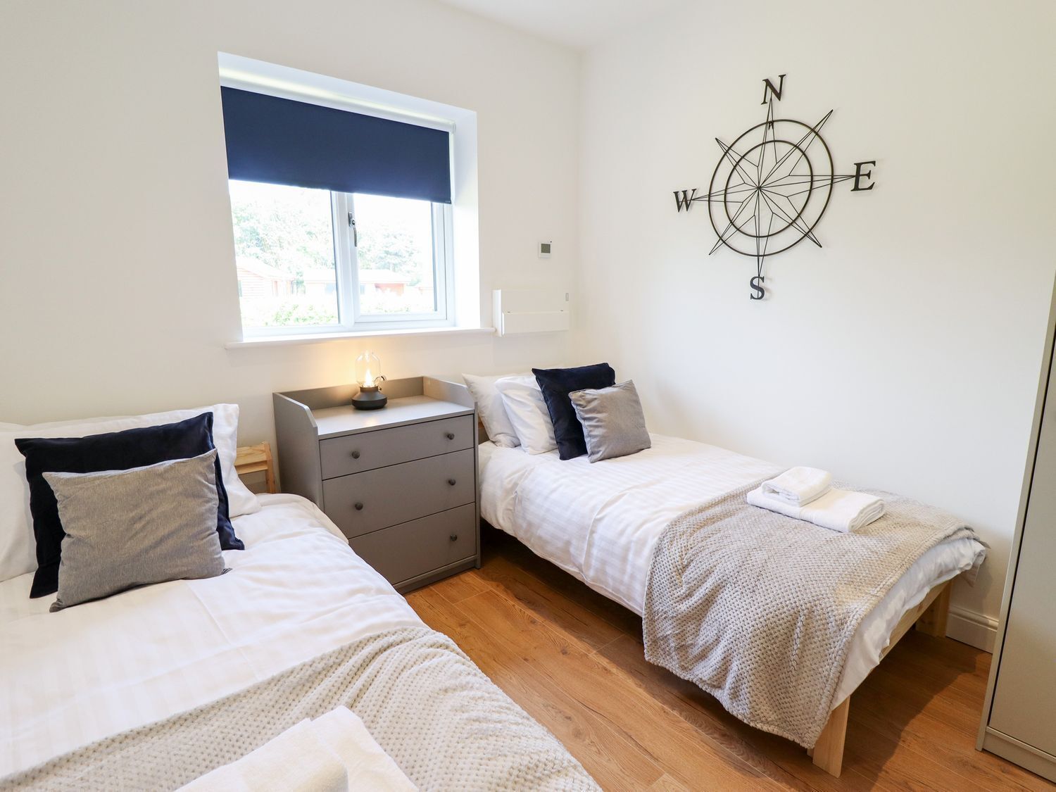 Een slaapkamer met twee bedden, een nachtkastje, een kompasdecoratie aan de muur, en een raam met een donker blind.