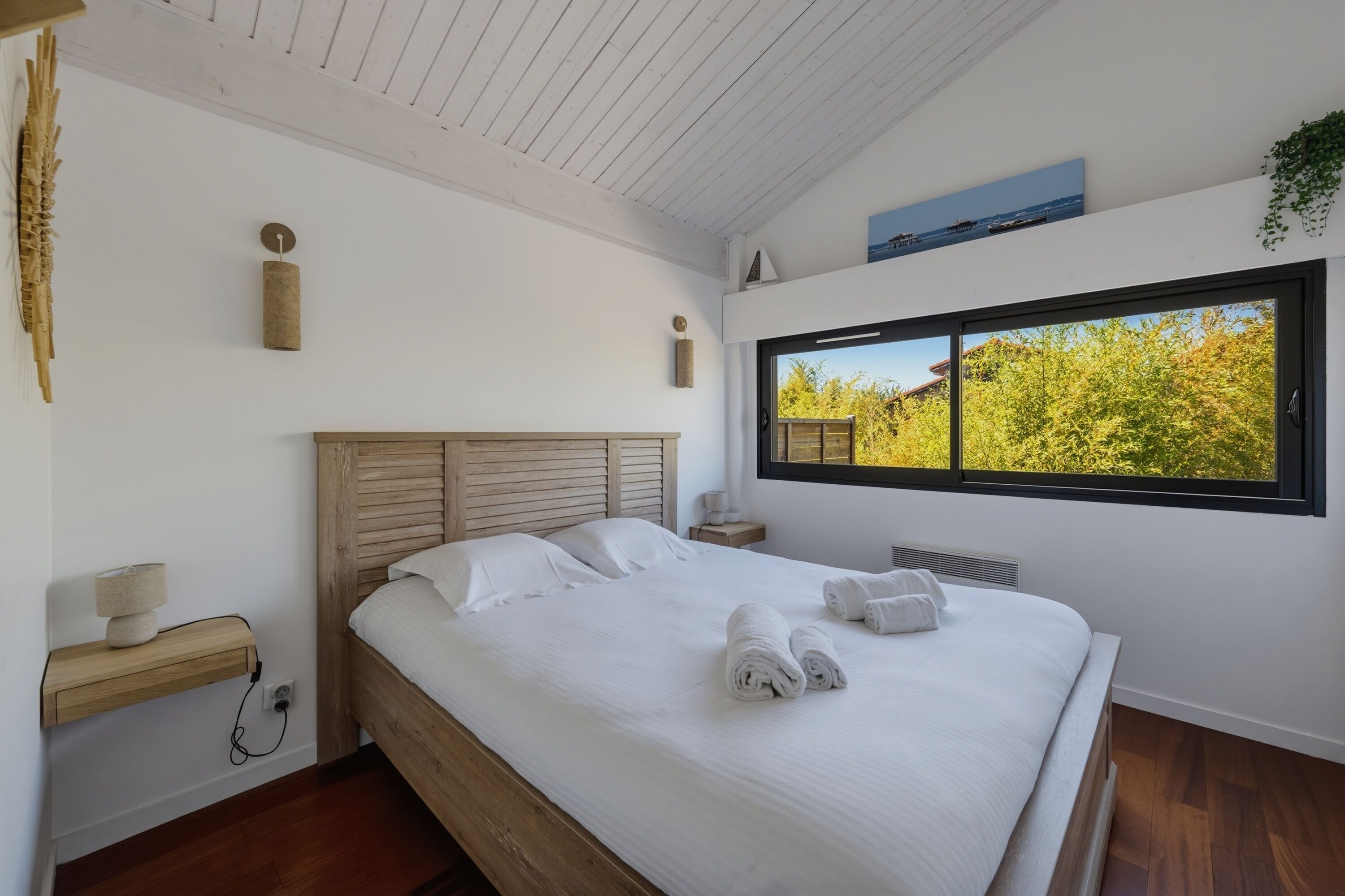 Ein Schlafzimmer mit einem hölzernen Bett, weißen Leinentüchern, einem Nachttisch, einer Lampe, Wanddeko und einem Fenster mit Blick auf Bäume.