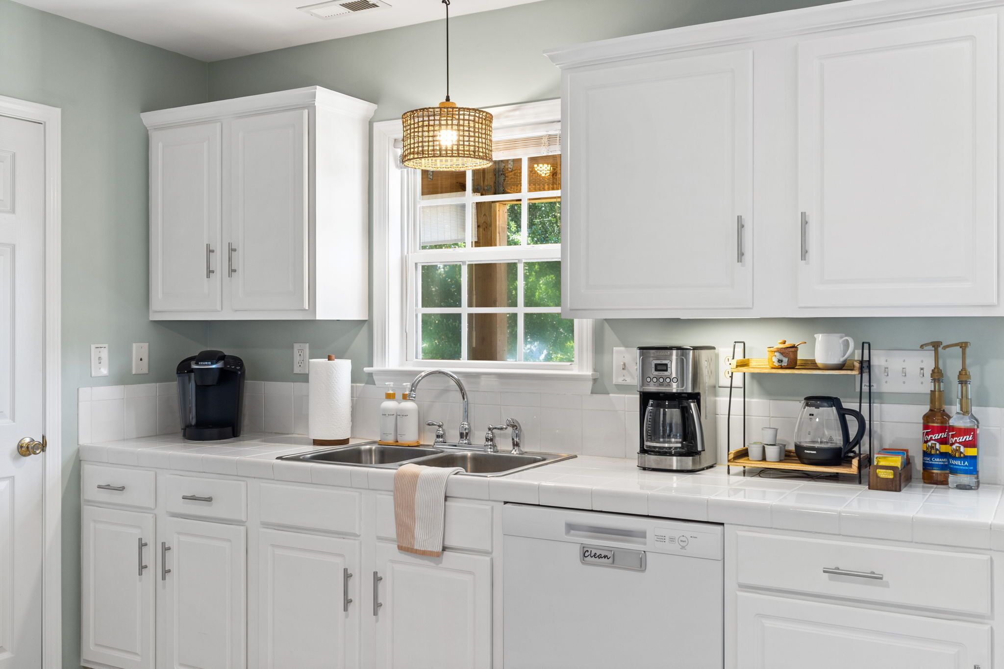 A kitchen with white cabinets, a sink, a coffee maker, and a window with a view of trees.