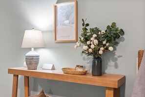 A wooden console table with a lamp, a framed picture, a vase with flowers, and a basket with jars.