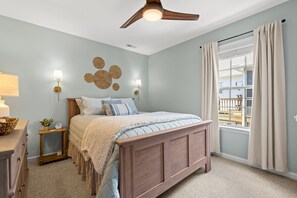 A bedroom with a bed, bedside lamps, a ceiling fan, a window with curtains, and a wall decor piece.