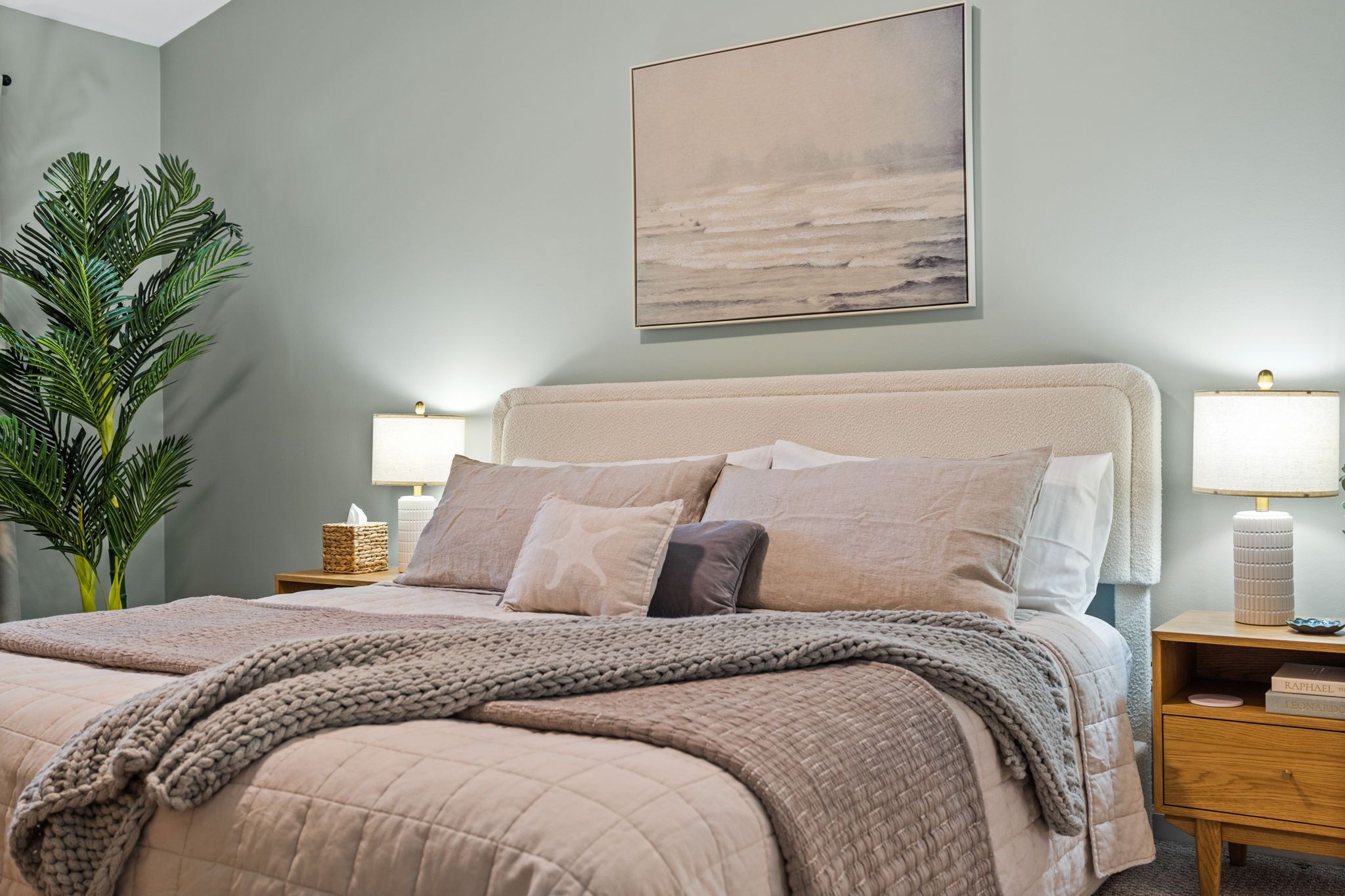 Een slaapkamer met een bed, nachtkastjes, een lamp, een plant en een schilderij aan de muur.