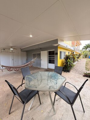 Une terrasse extérieure avec une table en verre et quatre chaises, un hamac et un bâtiment doté d’un balcon.