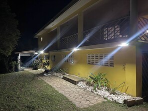 A two-story building with a yellow facade, a covered patio area, and a paved walkway with plants.