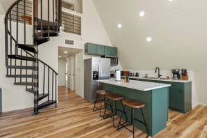 Eine moderne Küche mit einer zentralen Kochinsel, Barhocker, einem Edelstahlkühlschrank und einer Treppe mit Metallgeländer.