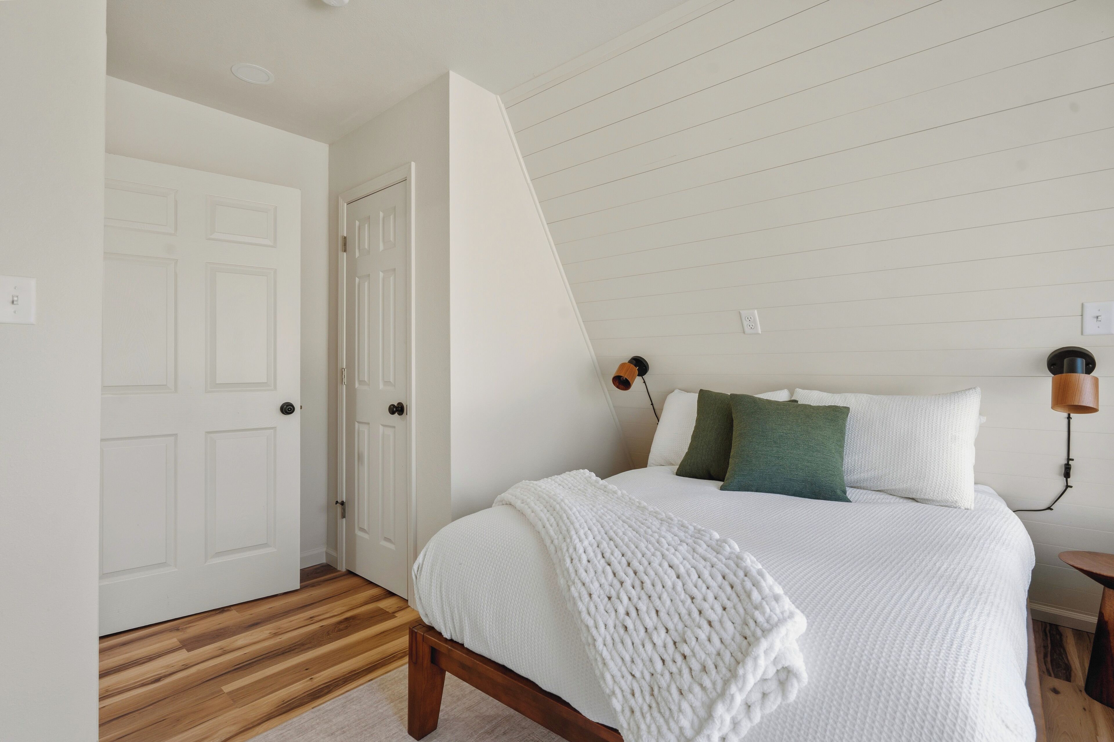 A bedroom with a wooden floor, a white bed with a white blanket, a green pillow, and a wooden nightstand with a lamp.
