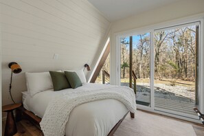 Une chambre à coucher comprenant un lit, une table de chevet, une lampe et une vue sur une zone boisée, visible à travers une porte-fenêtre coulissante.