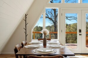 Une table à manger dressée avec des assiettes, un vase et un porte-manteau, dans une pièce dotée d’un plafond en pente et de grandes fenêtres.