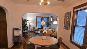 Ein Essbereich mit einem runden Tisch, vier Stühlen, einem Deckenventilator und Blick in ein Wohnzimmer mit einer Couch und einer Topfpflanze.