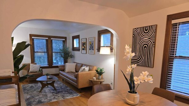 Ein Wohnzimmer mit einer Couch, einem Couchtisch, einem Esstisch und Stühlen. An den Wänden hängen Bilderrahmen, eine Lampe steht dort, und es gibt einen Blumentopf.