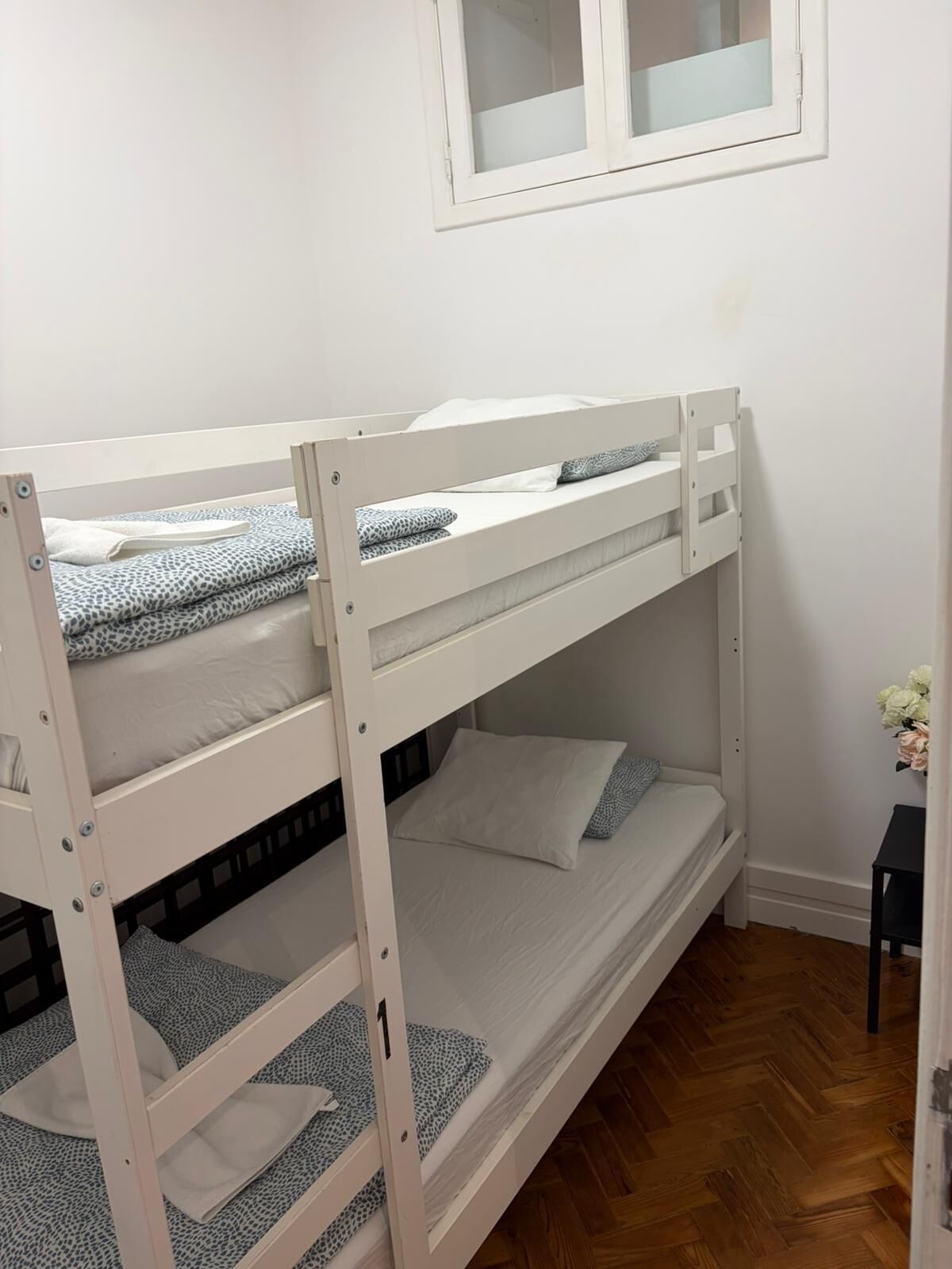 Dormitorio con literas, ventana, suelo de madera y una pequeña mesa con flores.