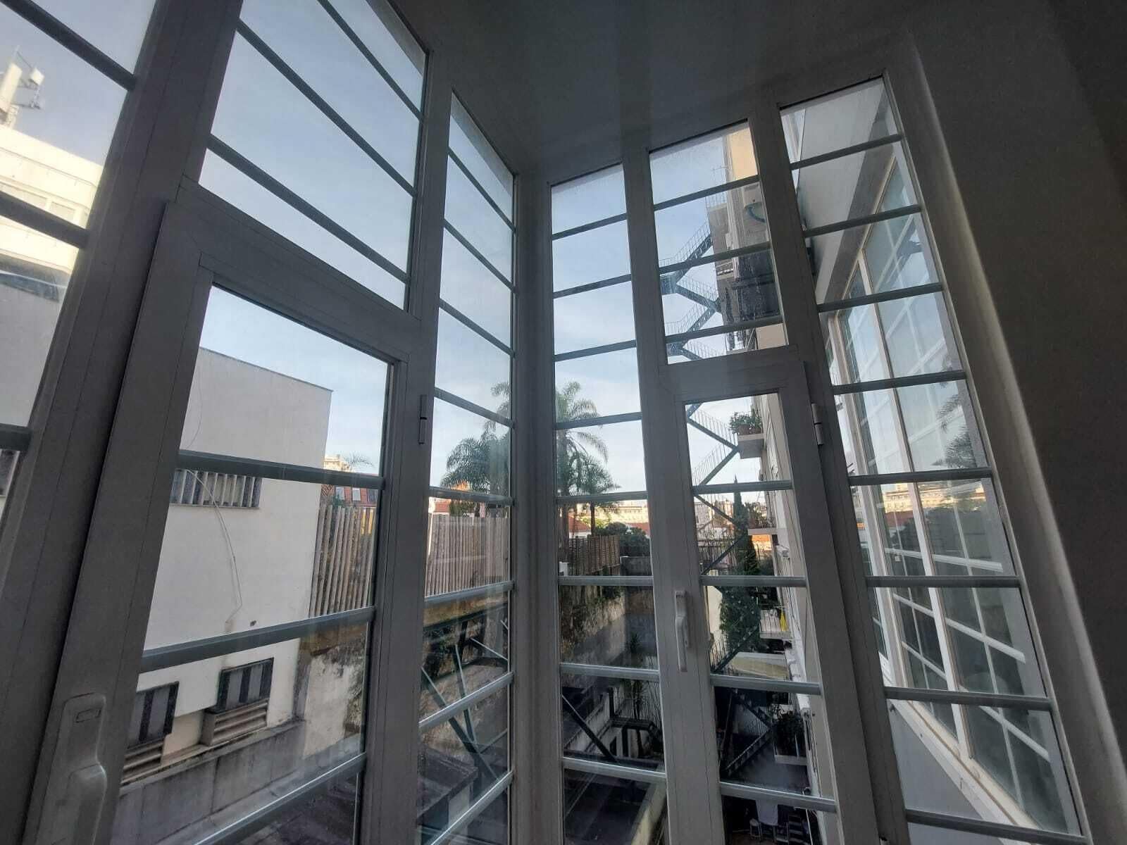 Huoneen nurkka, jossa on suuret ikkunat, näkyviin jäävät portaat ja näkymä naapurirakennuksiin.