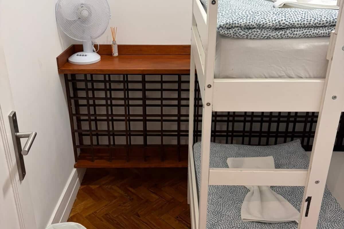 A bunk bed with patterned bedding, a white fan on a wooden table, a trash bin, and a wooden floor.