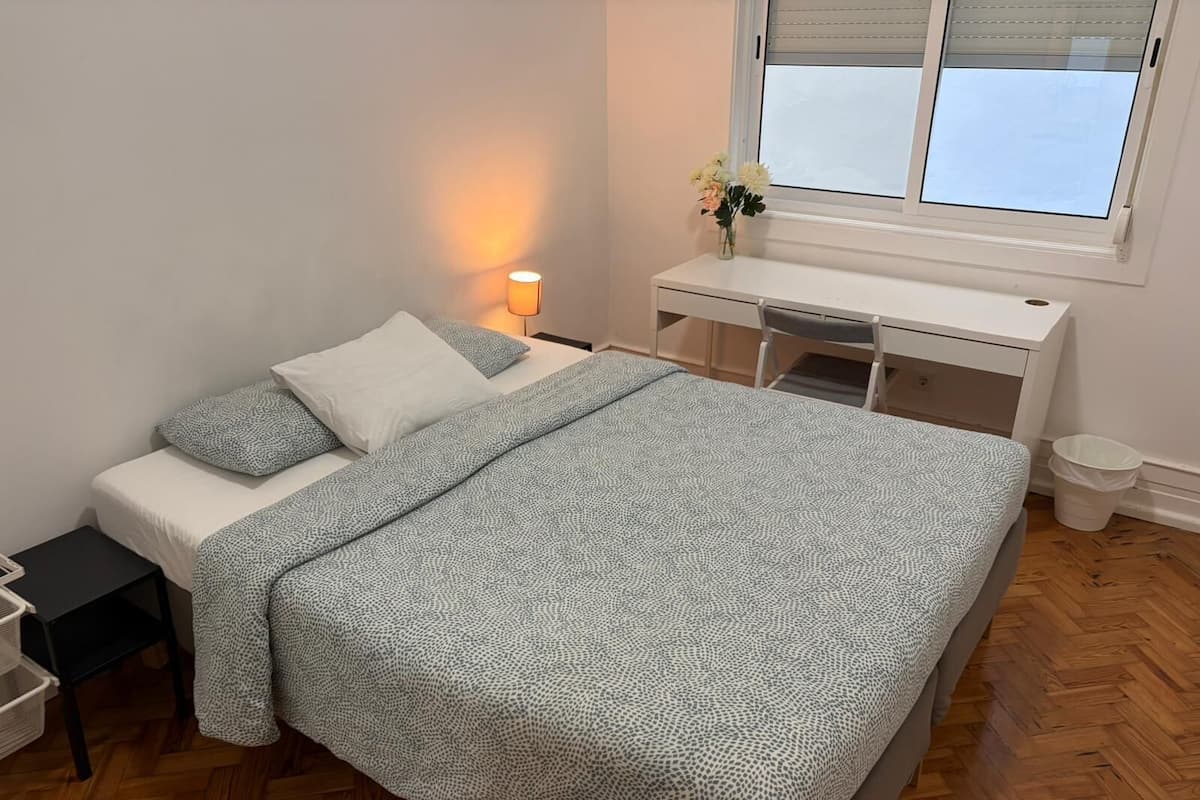 A neatly arranged bedroom with a bed, bedside table, desk, and a window with blinds.