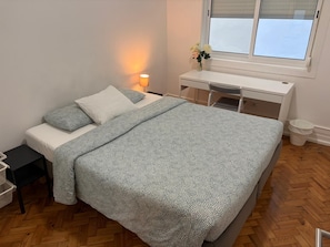 A neatly arranged bedroom with a bed, bedside table, desk, and a window with blinds.